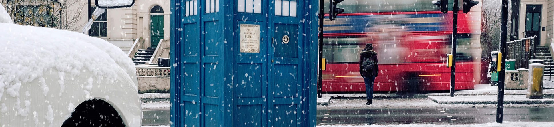 Tardis on snow covered pavement next to pelican crossing with pedestrian walking away over the crossing, pausing as a red double-decker bus passes. On left in foreground is the front right wheel an arch of a white car.