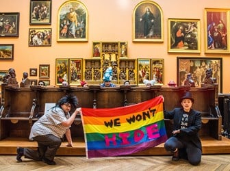 9 drag performers of various ages, genders and ethnicities pose in a large museum room with religious paintings on the walls. 7 of the performers are hiding behind an ornate wooden structure in the centre of the room, with only their heads showing. 2 of the performers keel in front of the structure holding a rainbow flag into which the following text has been sewn in black and pink lettering "We Won't Hide"