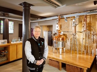 A short-haired lady standing with her hands clasped in front of a gin distillery. 