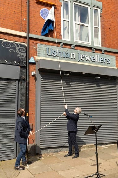 Two men in suits unveiling a blue heritage plaque set in a red brick wall building with grey shop shutters