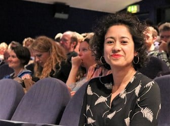 A portrait photograph of Samira Ahmed sitting in a blue auditorium seat, with other people sitting in the seats behind her