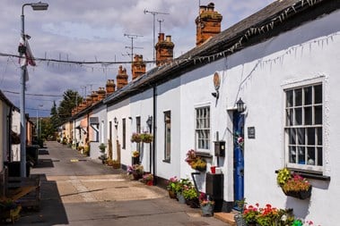 A row of colourful single-storey cottages, with flowers outside, and fairy lights hanging along a narrow street.