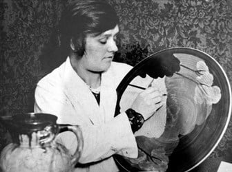 A black and white photograph of a woman in a white coat concentrating while painting a decorative ceramic plate