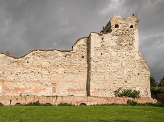 The ruined corner of a large stone castle, several storeys high, in parkland, with stormy skies overhead. 