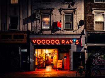 A photograph of a restaurant at night. The interior is lit yellow and the sign above reads 'Voodoo Ray's' in red light. 