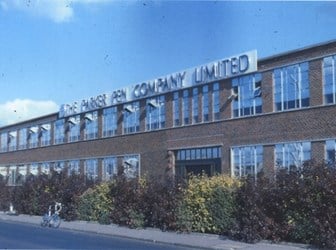 A photograph of a factory building with many glass windows. A sign at the top of the building reads "THE PARKER PEN COMPANY LIMITED".