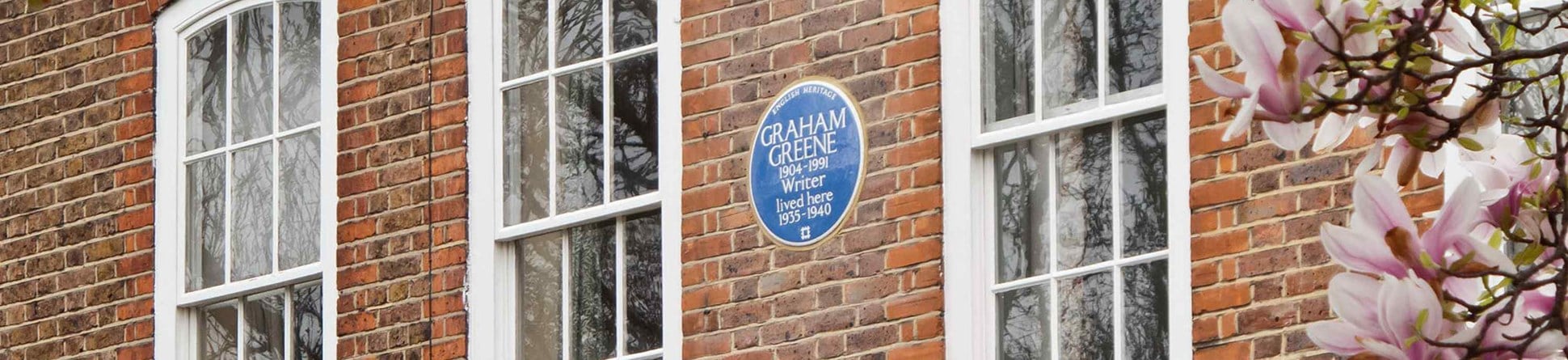 Close-up of a blue heritage plaque between large windows on a red brick wall. Tree foliage frames the image on the far left and far right.