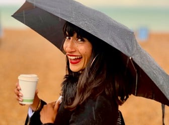A portrait photograph of Susie Thornberry on a beach smiling while holding an umbrella and a takeaway coffee cup