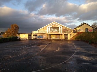 A building with graffiti in the evening light. An empty tarmac car park is in the foreground.